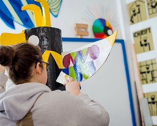 Eine Person bemalt ein großes, buntes Kunstobjekt in einem Atelier.